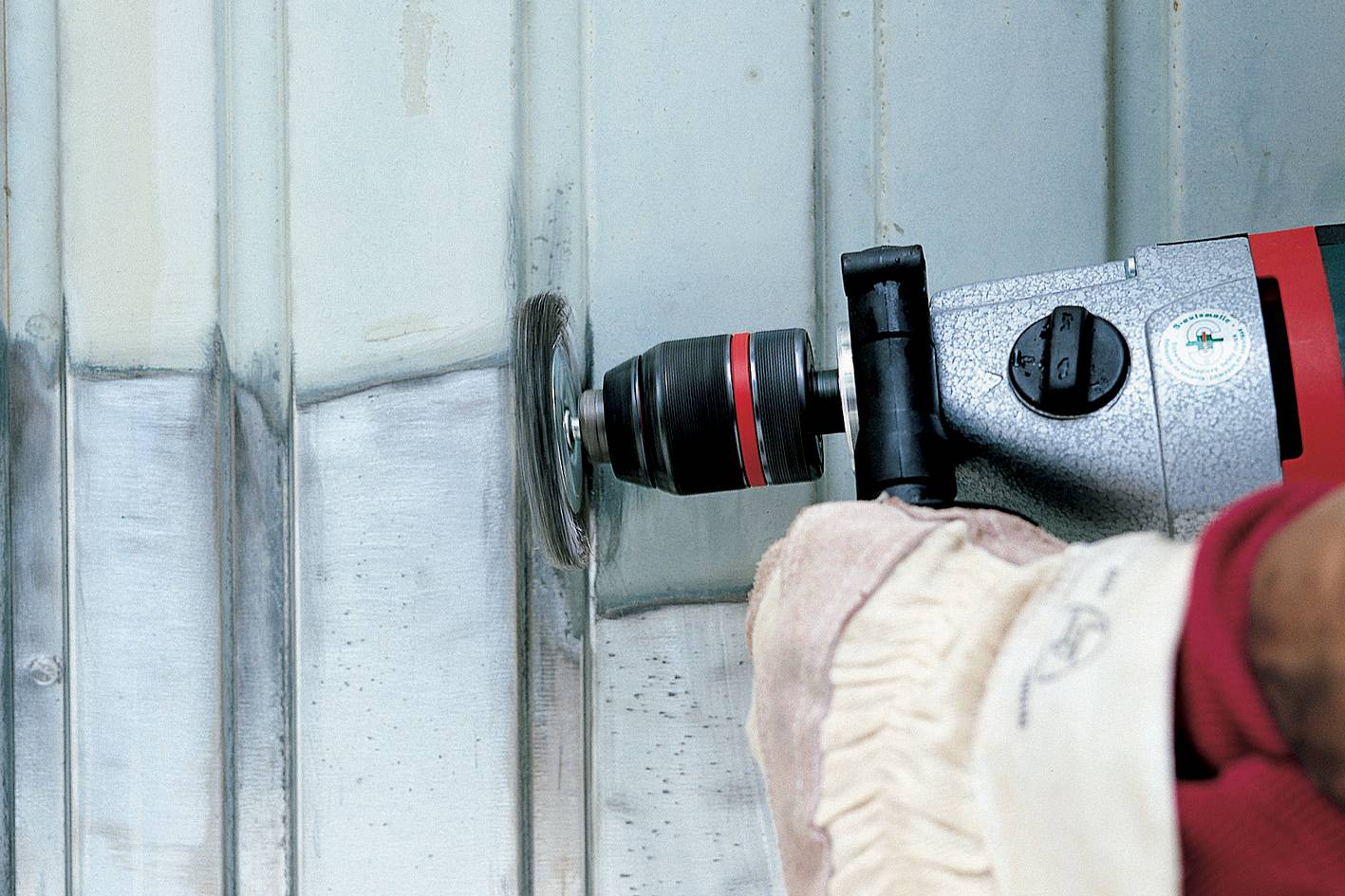 Osoba čistí kovovú plochu elektrickým náradím. Pravá ruka drží náradie, kým ľavá ruka pridržiava kov.