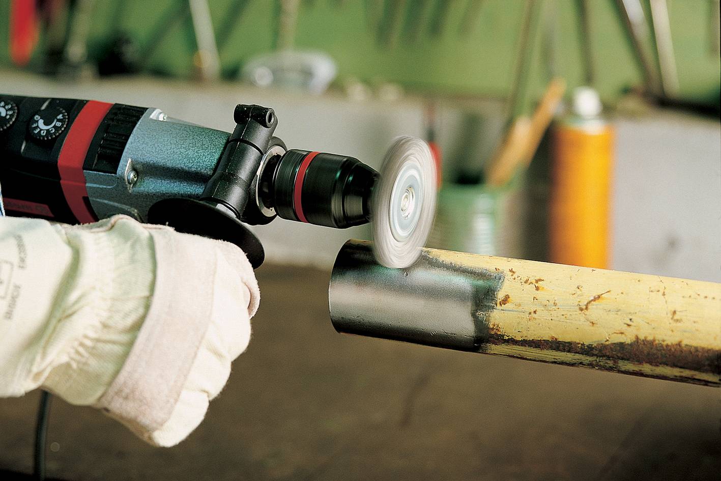 Osoba brúsi kovové potrubie elektrickým brúsnym strojom, aby vyhladila povrch. V pozadí je dielňa.