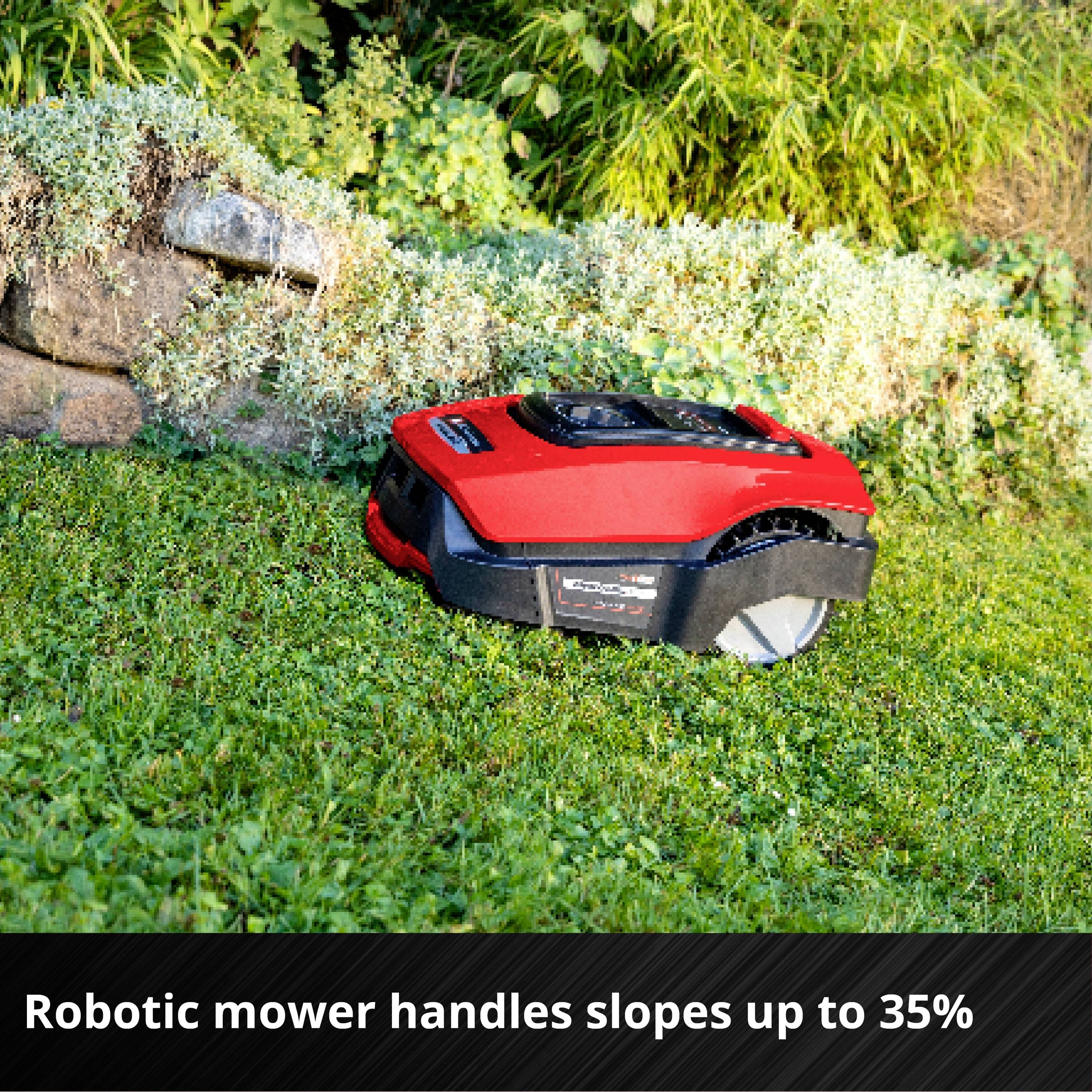 Červený robotický kosačkár samostatne kosí trávu na zelenej trávnatej ploche vedľa kamennej steny. Text: 'Zvládne svahy so sklonom až do 35% bez problémov'.