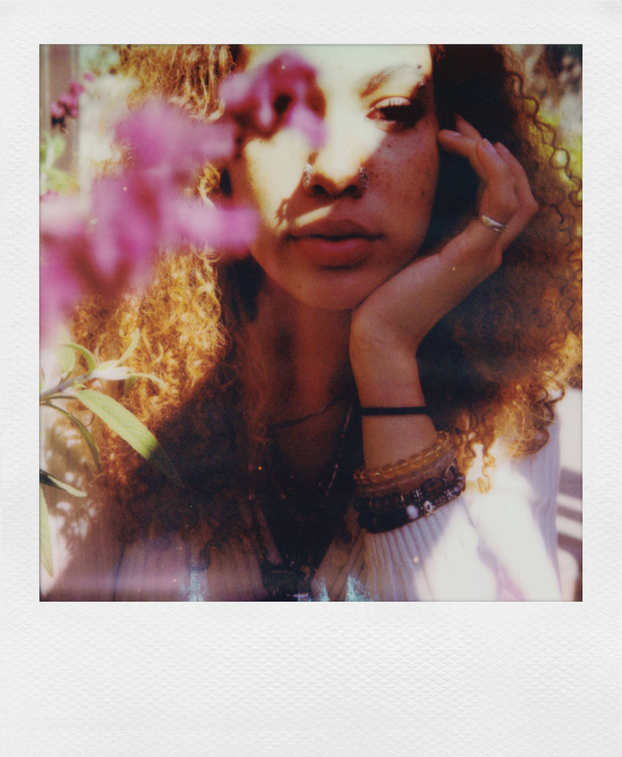 Osoba s kučeravými vlasmi si podopiera bradu rukou a díva sa priamo do kamery. V popredí sú rozmazané kvety.