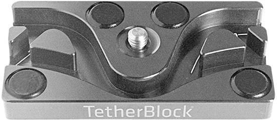 Kovový adaptér na vedenie kábla so skrutkou v strede. Názov produktu 'TetherBlock' je na prednej strane.