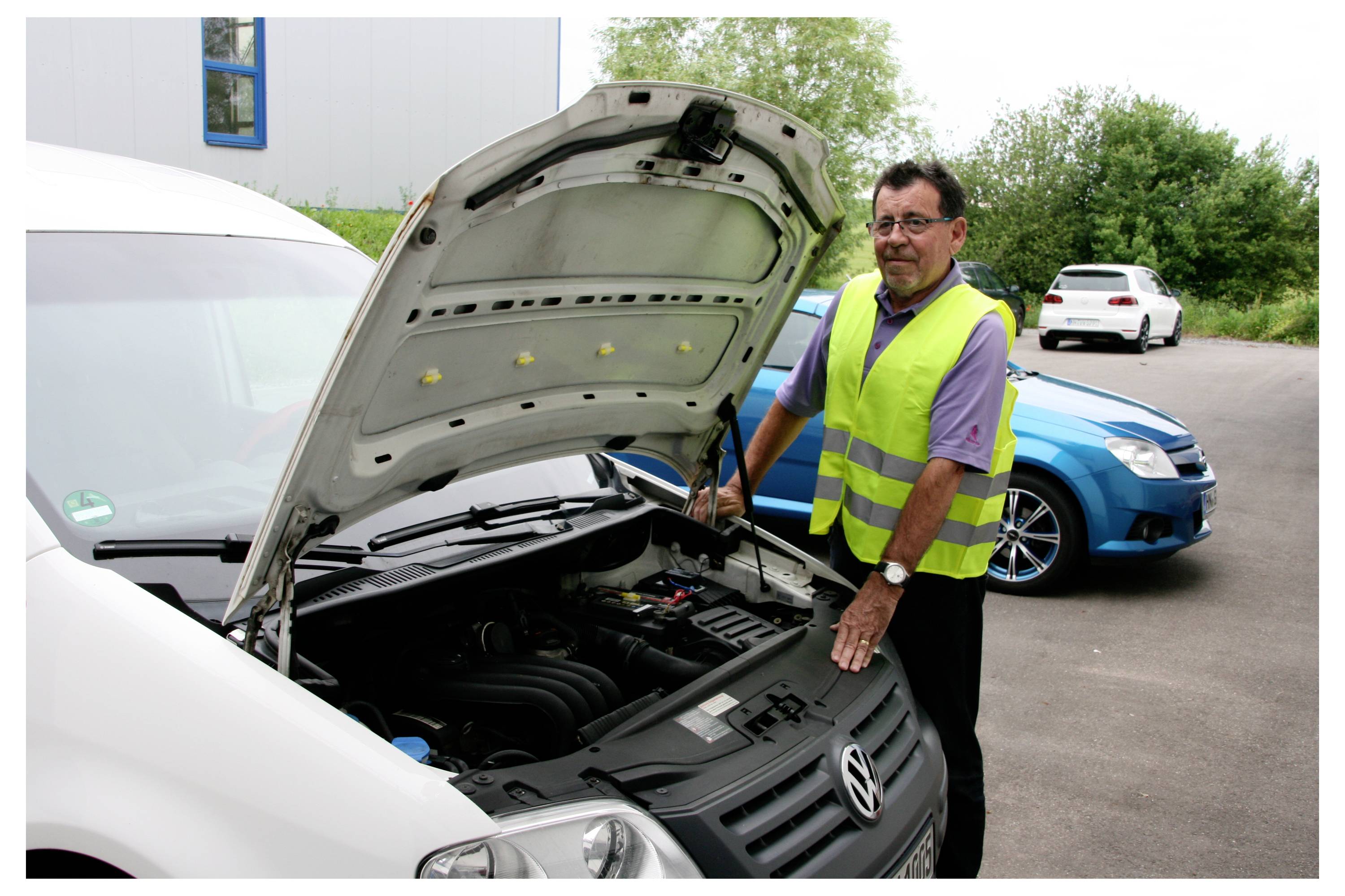 Osoba v fluorescenčnej veste kontroluje motor bieleho dodávkového vozidla s otvoreným motorovým priestorom. Neďaleko je zaparkovaný modrý automobil.