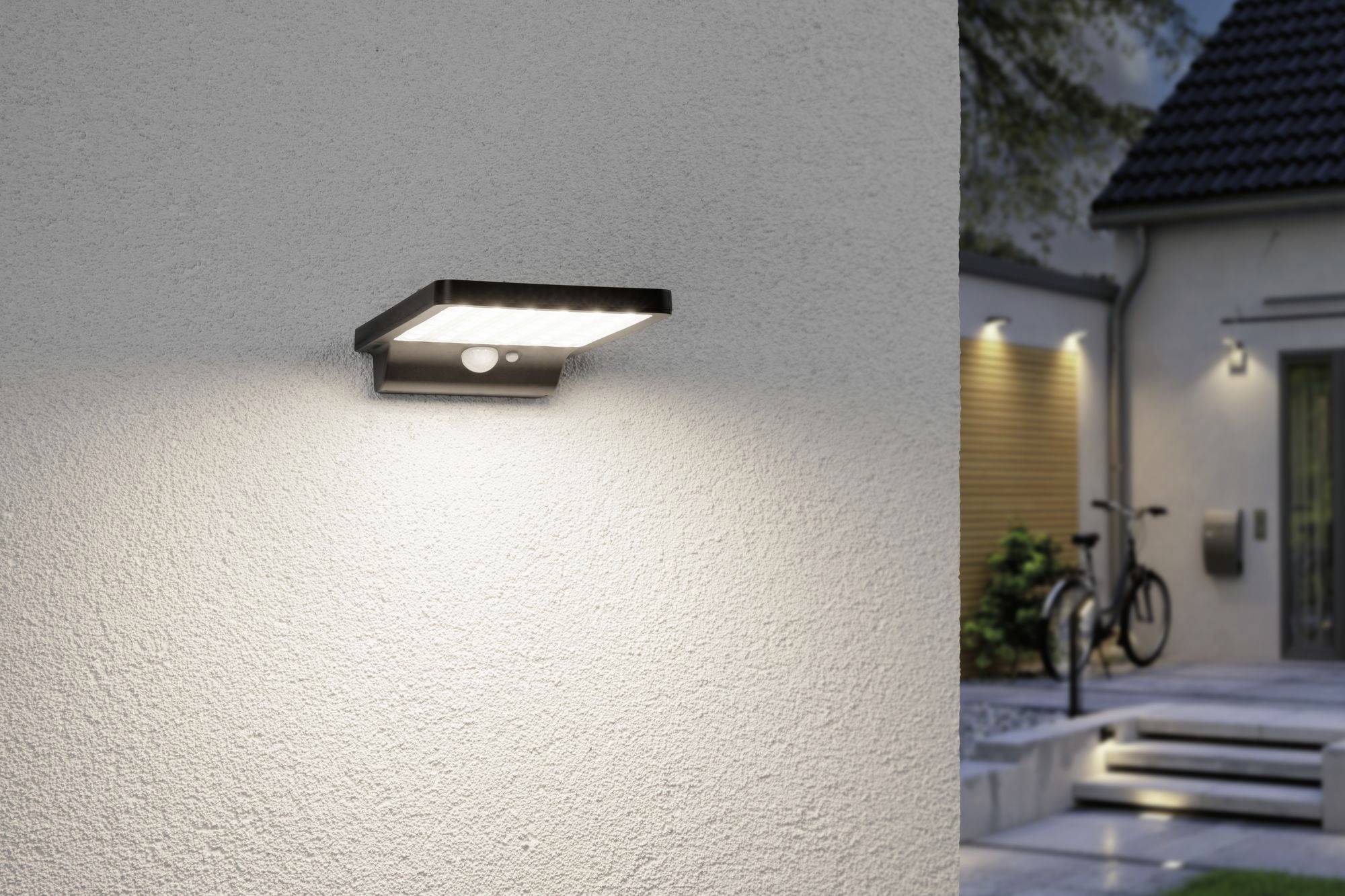 Nástenné svietidlo s pohybovým senzorom na stene domu osvetľuje cestu k modernému vstupu za súmraku. V pozadí je bicykel.
