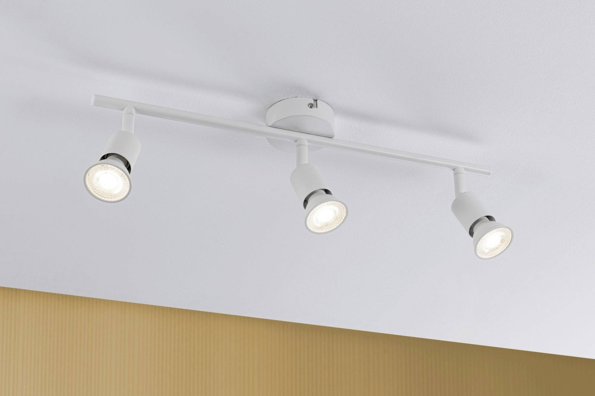 Tri biele stropné svetlá svietia, upevnené na bielom držiaku pred béžovou stenou.