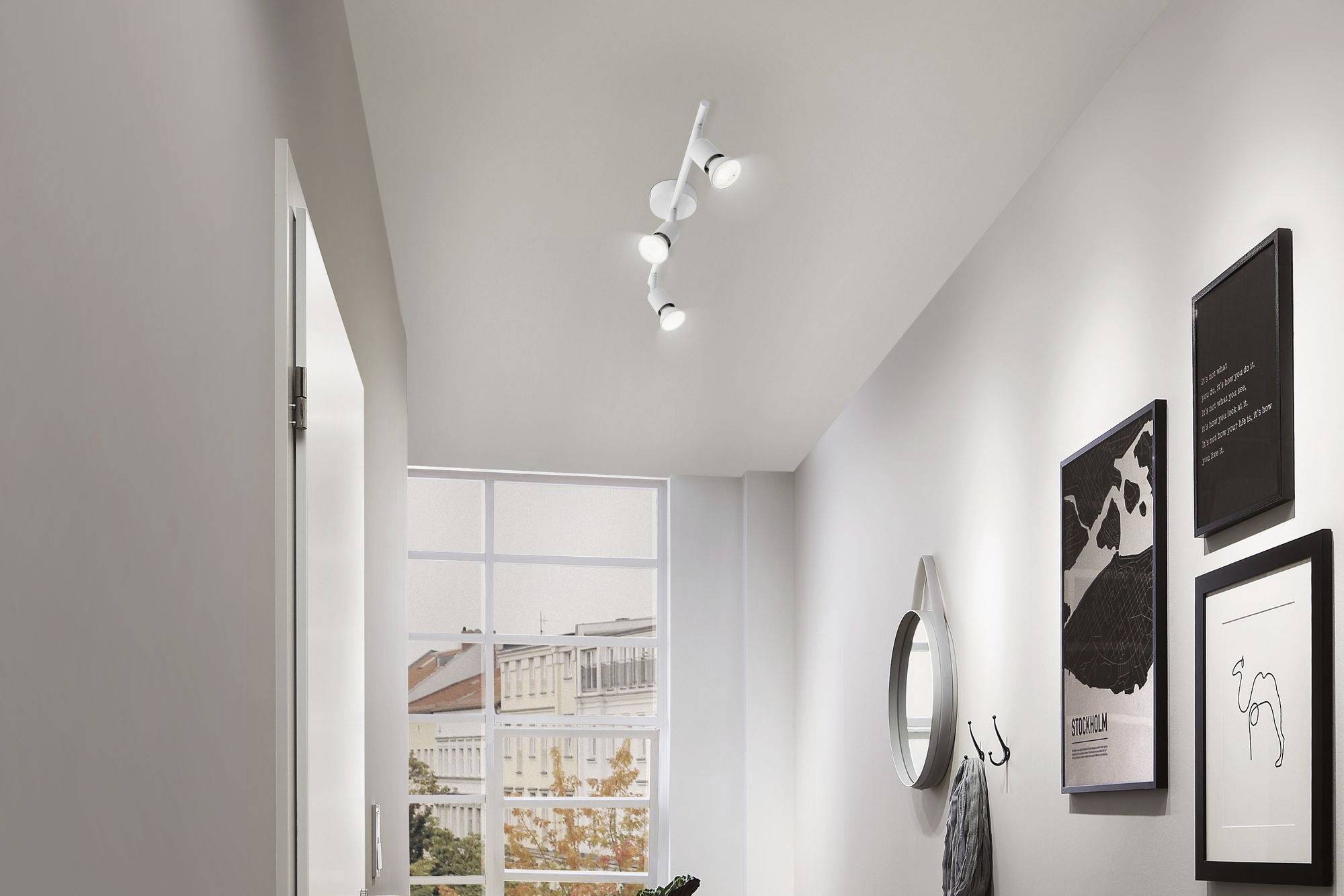 Chodba s bielym stropom a stenou, na ktorej visia zarámované obrazy. Moderný luster osvetľuje priestor, okno ponúka výhľad von.