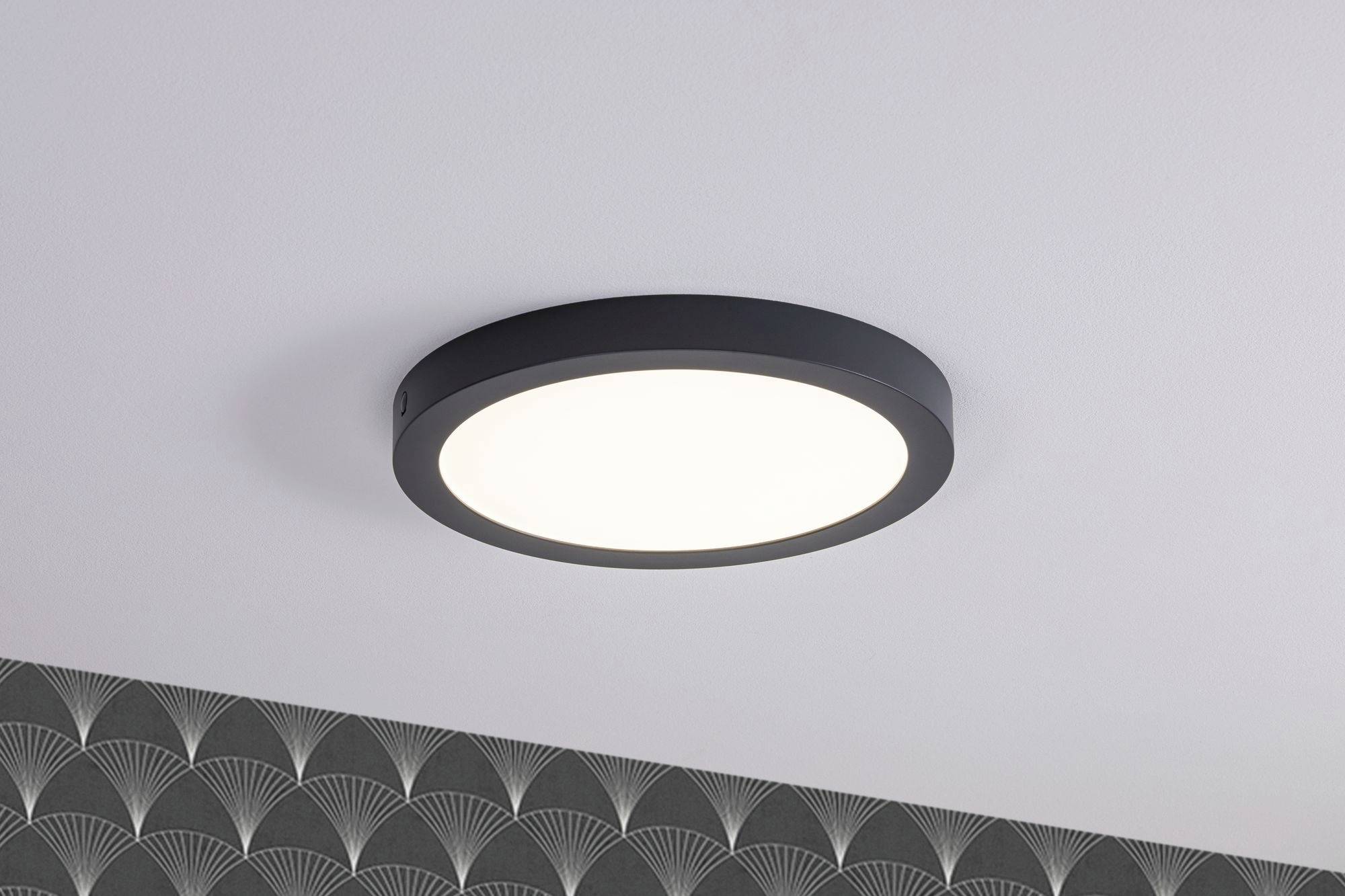 Okrúhle stropné svietidlo s čiernym rámom na bielom strope, vyžarujúce jasné svetlo; pod ním tapeta s vejárovým dizajnom.