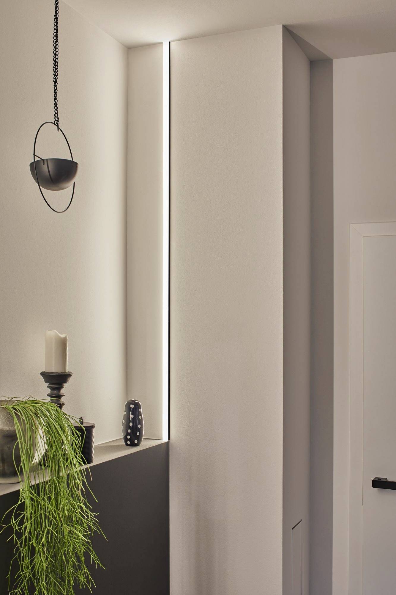 Minimalistický, moderný interiér s úzkym svetelným pruhom na stene, jednoduchým svietníkom, dekoratívnou vázou a visiacou rastlinou.