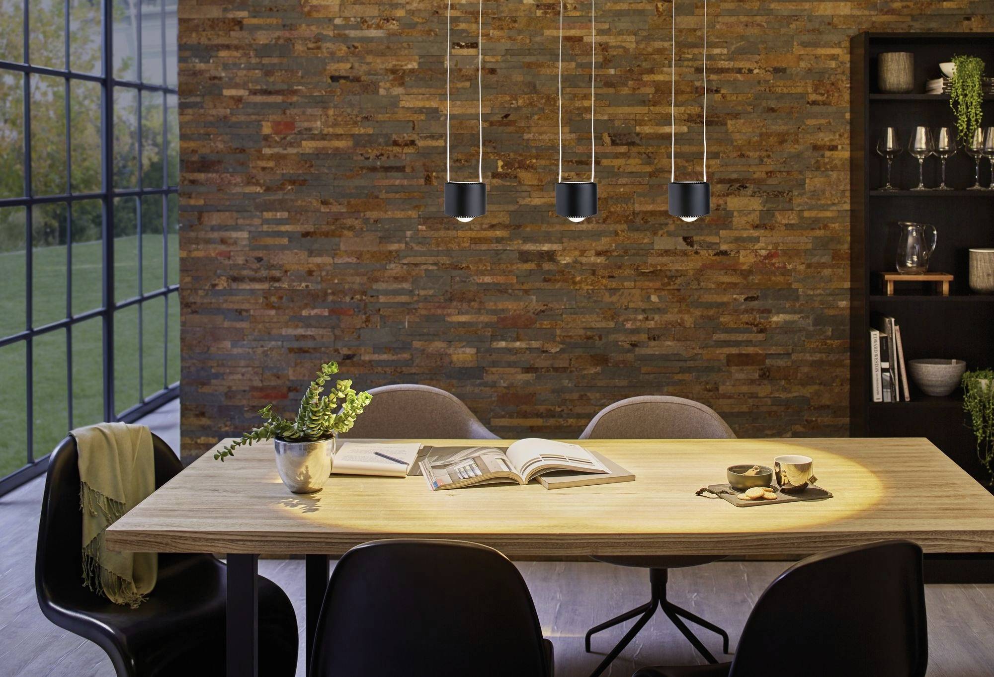 Moderne zariadený jedálenský priestor s dreveným stolom, stoličkami, visiacimi lampami a kamennou stenou. Na stole leží kniha a sú tam umiestnené rastliny.