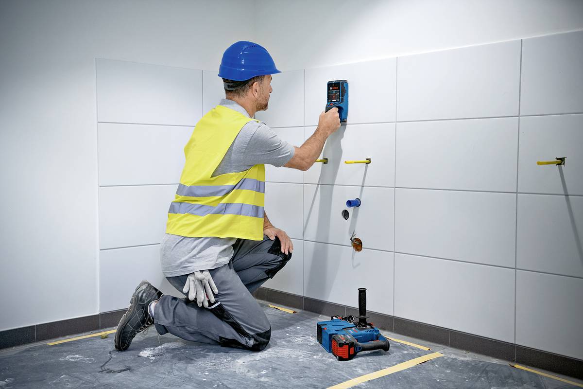 Stavebný robotník kľačí na zemi a pomocou elektronického zariadenia meria vlhkosť obkladačkovej steny. Má na sebe žltú bezpečnostnú vestu a modrú prilbu.