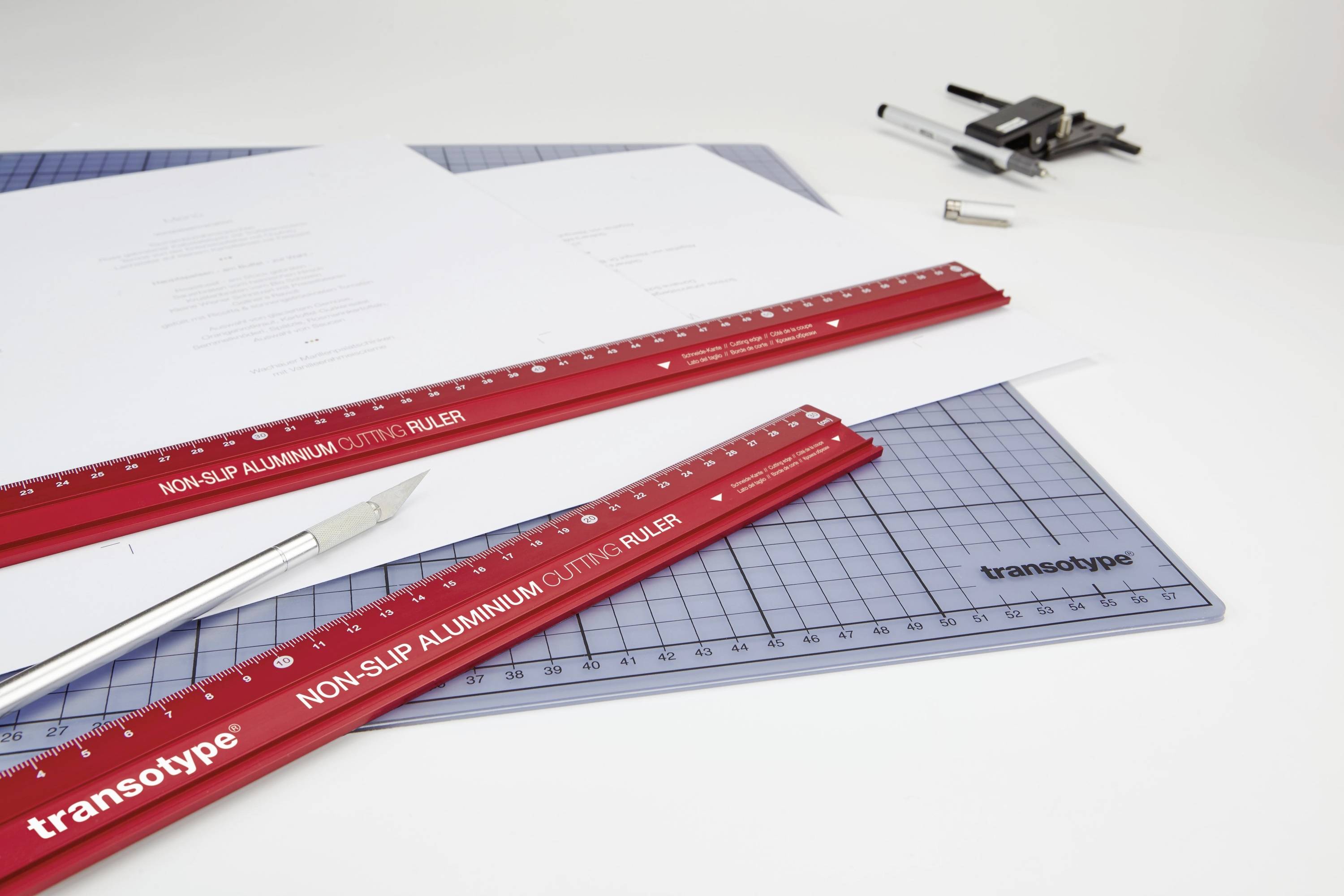 Červené hliníkové pravítko s nápisom 'NON-SLIP ALUMINIUM CUTTING RULER' leží na rezacej doske. Vedľa neho je odlamovací nôž a perá.