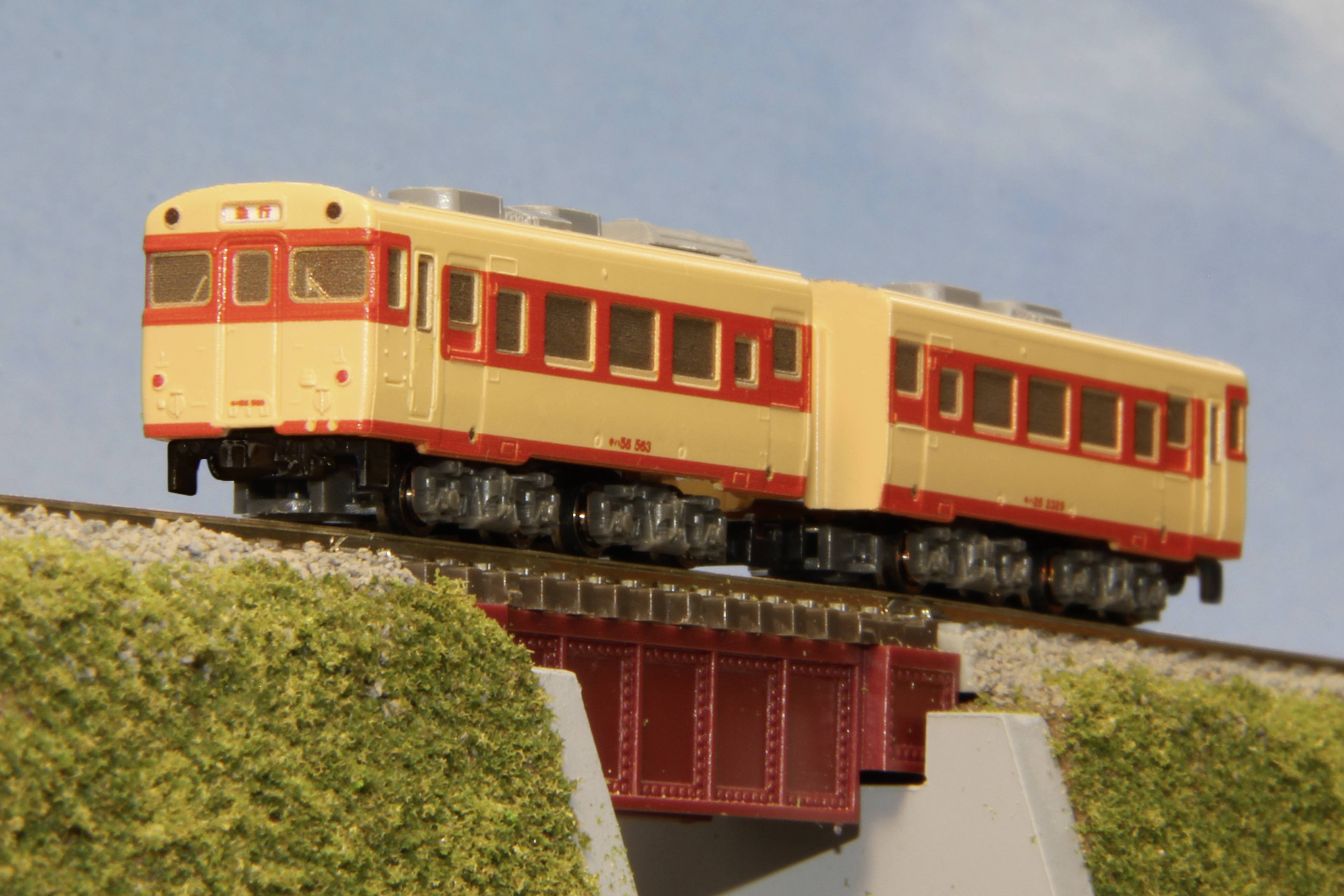 Žltý a červený modelový vlak jazdí po železničnom moste cez zatrávnený terén pod modrým nebom.