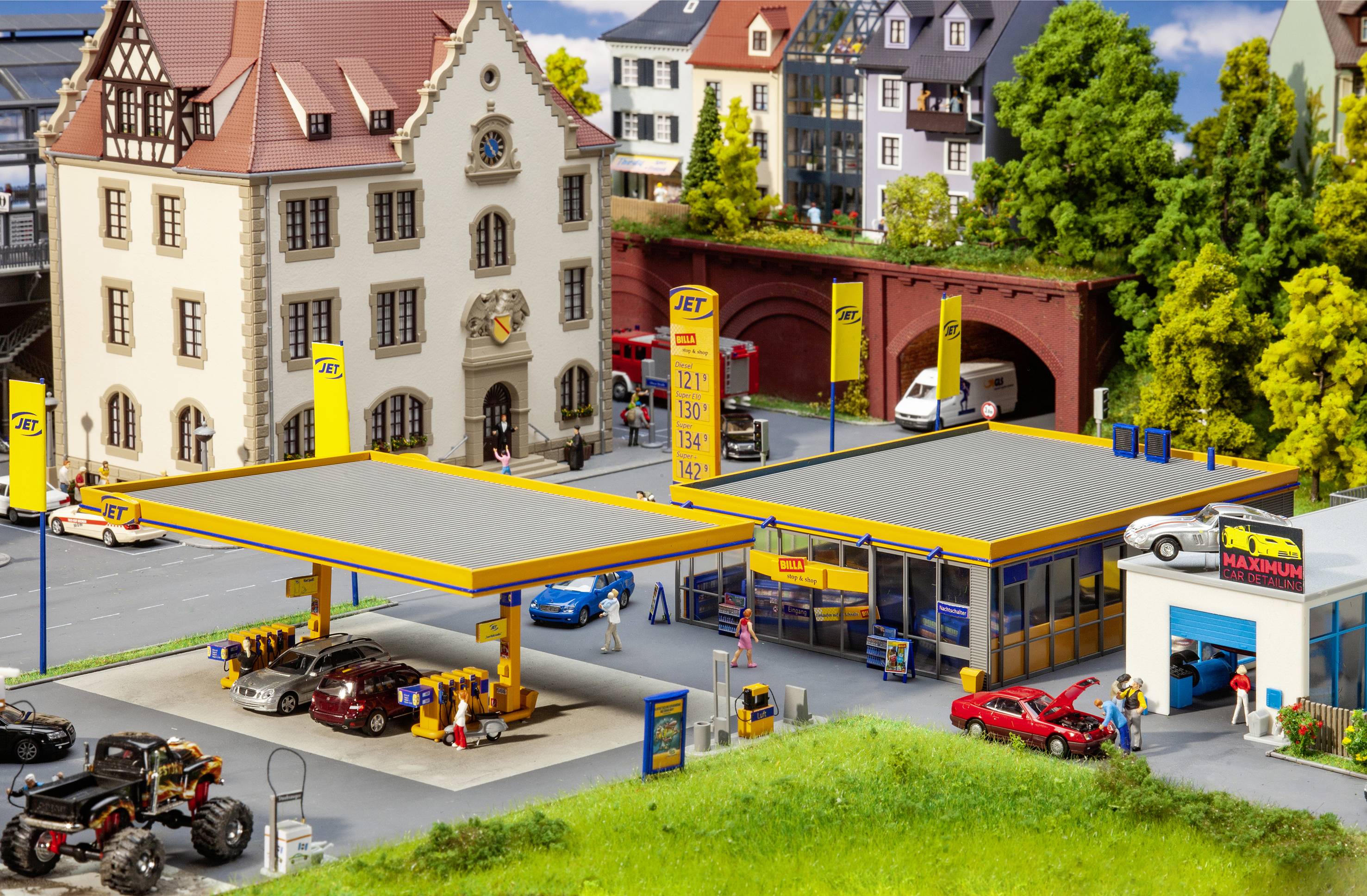 Miniaturový model čerpacej stanice JET s autami, obchodom a okolitými budovami. V pozadí je vidieť most a stromy.