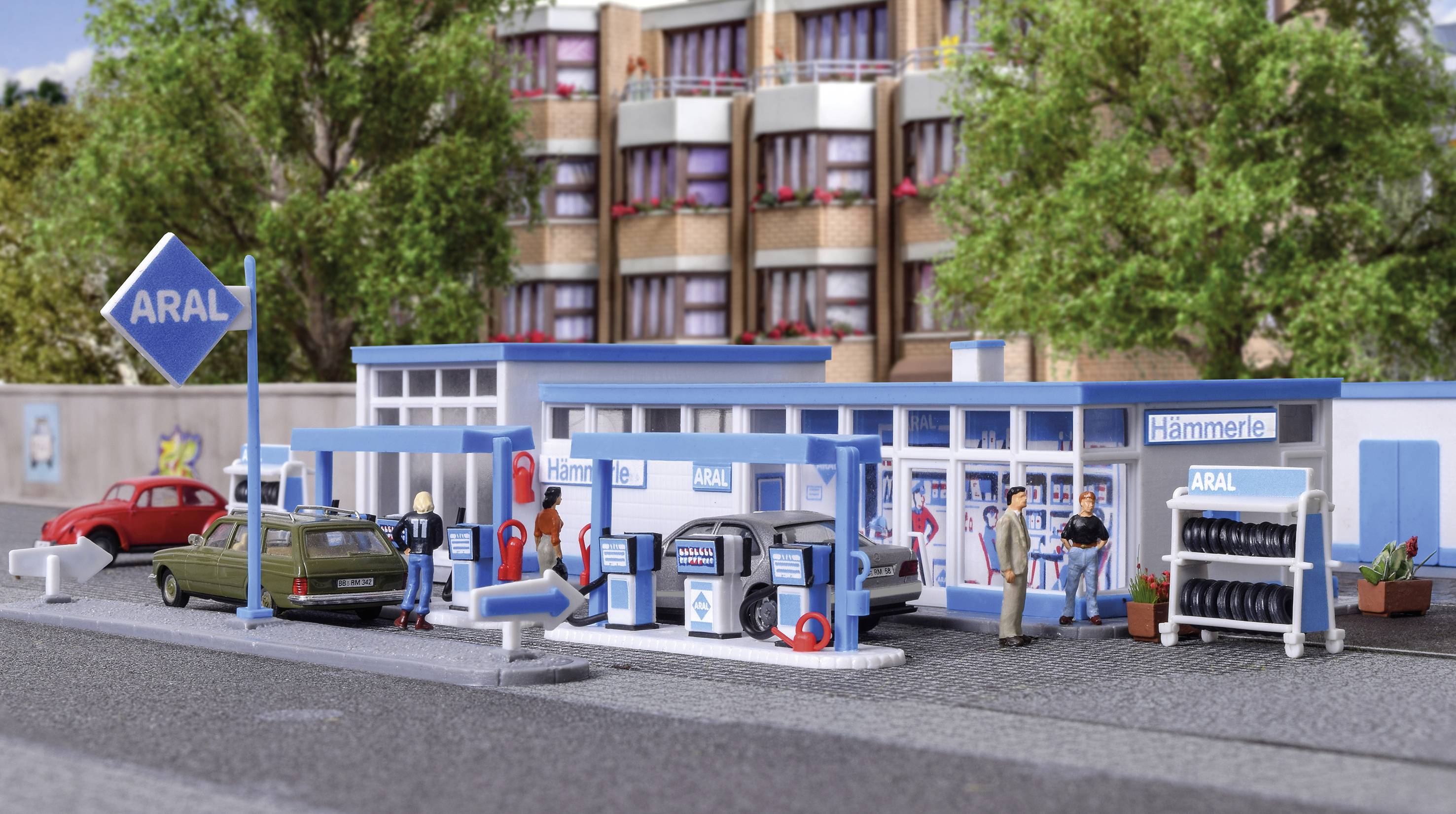 Miniaturná čerpacia stanica s logom 'Aral', niekoľkými stojanmi na čerpanie paliva, parkujúcimi autami a ľuďmi. V pozadí sú viditeľné budovy a stromy.