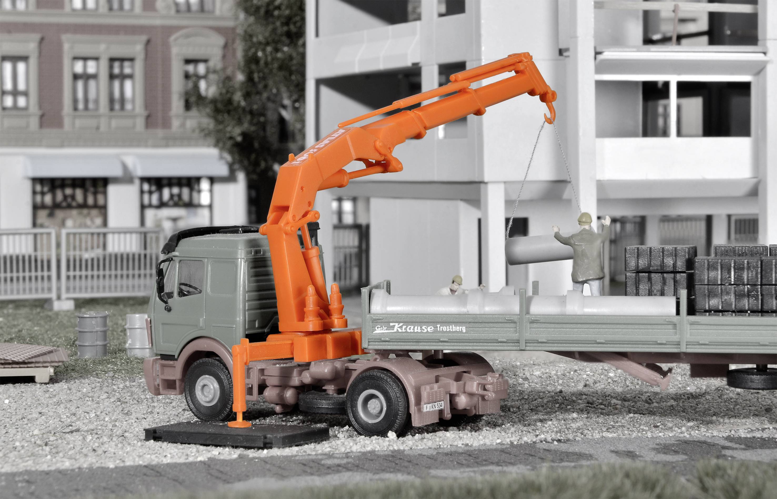 Nákladné auto s oranžovým žeriavom vykladá potrubia z budovy staveniska. V pozadí sú čiastočne postavené budovy.
