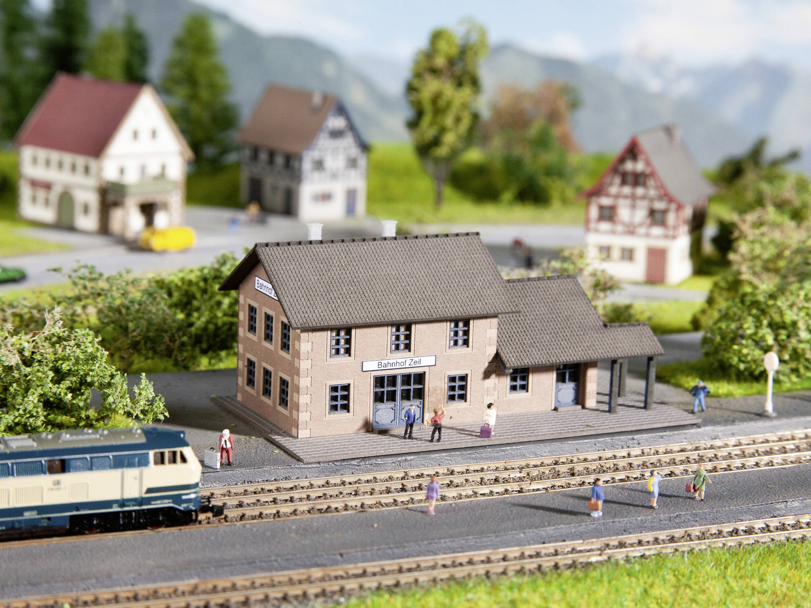 Miniatúrna železničná krajina zobrazuje vidiecku železničnú stanicu s prechádzajúcim vlakom, obklopenú zmenšenými domami a stromami.