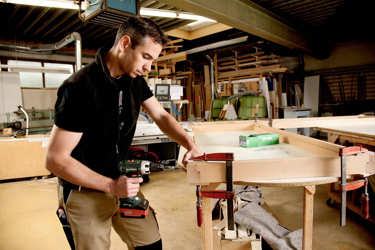 Muž v dielni používa vŕtačku na opracovanie dreva. V pozadí sú viditeľné rôzne nástroje a stroje.