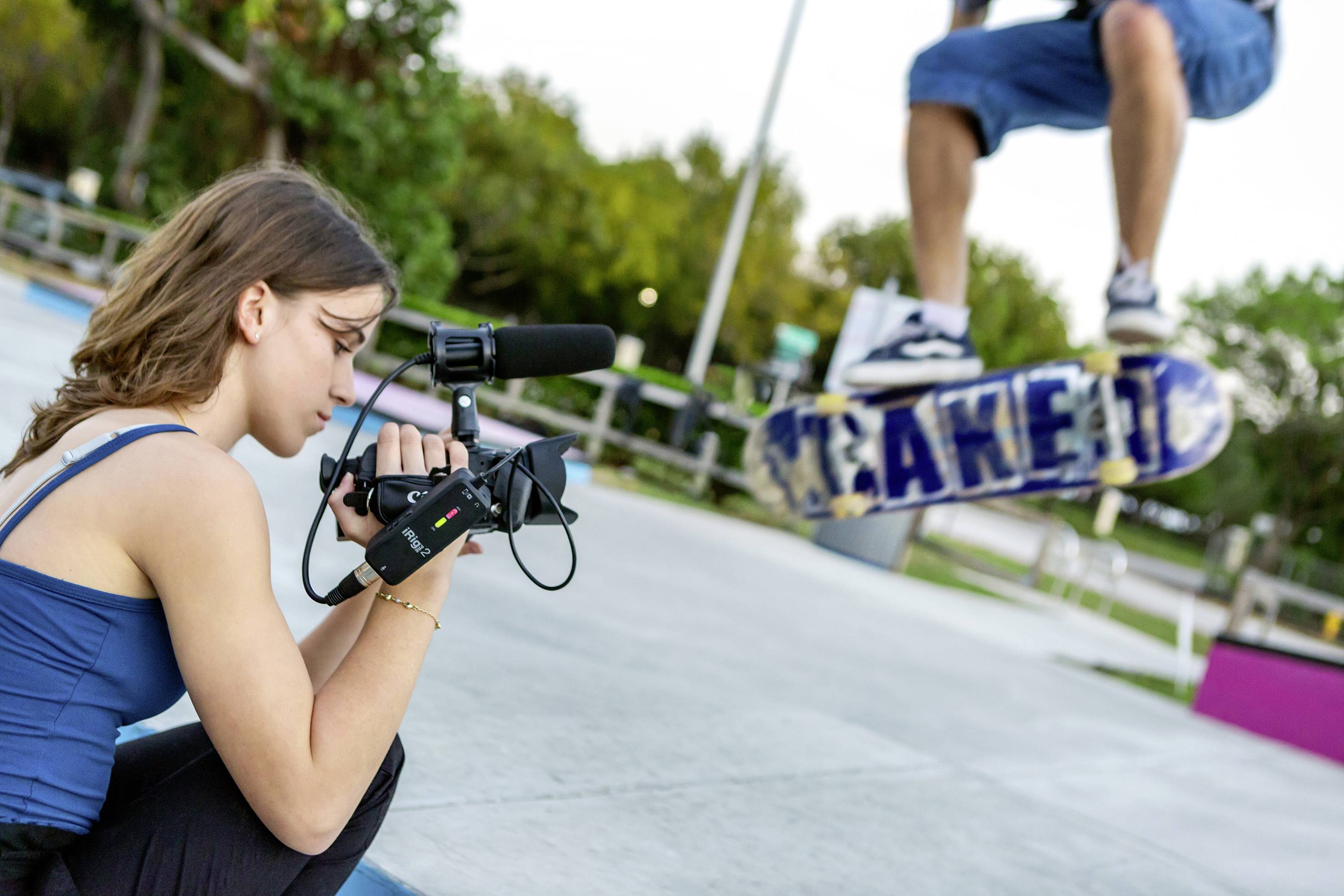 Mladá žena natáča videokamerou skateboardistu, ktorý skáče vo vzduchu. Okolie je skatepark.