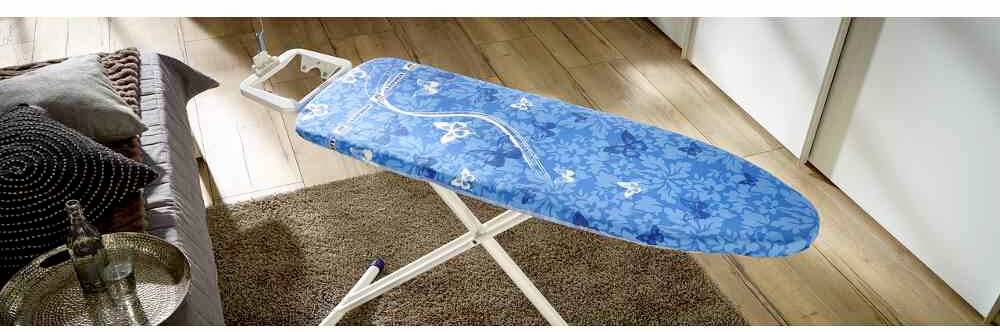 Žehliaca doska s modrým kvetinovým poťahom stojí na koberci v obytnej miestnosti.