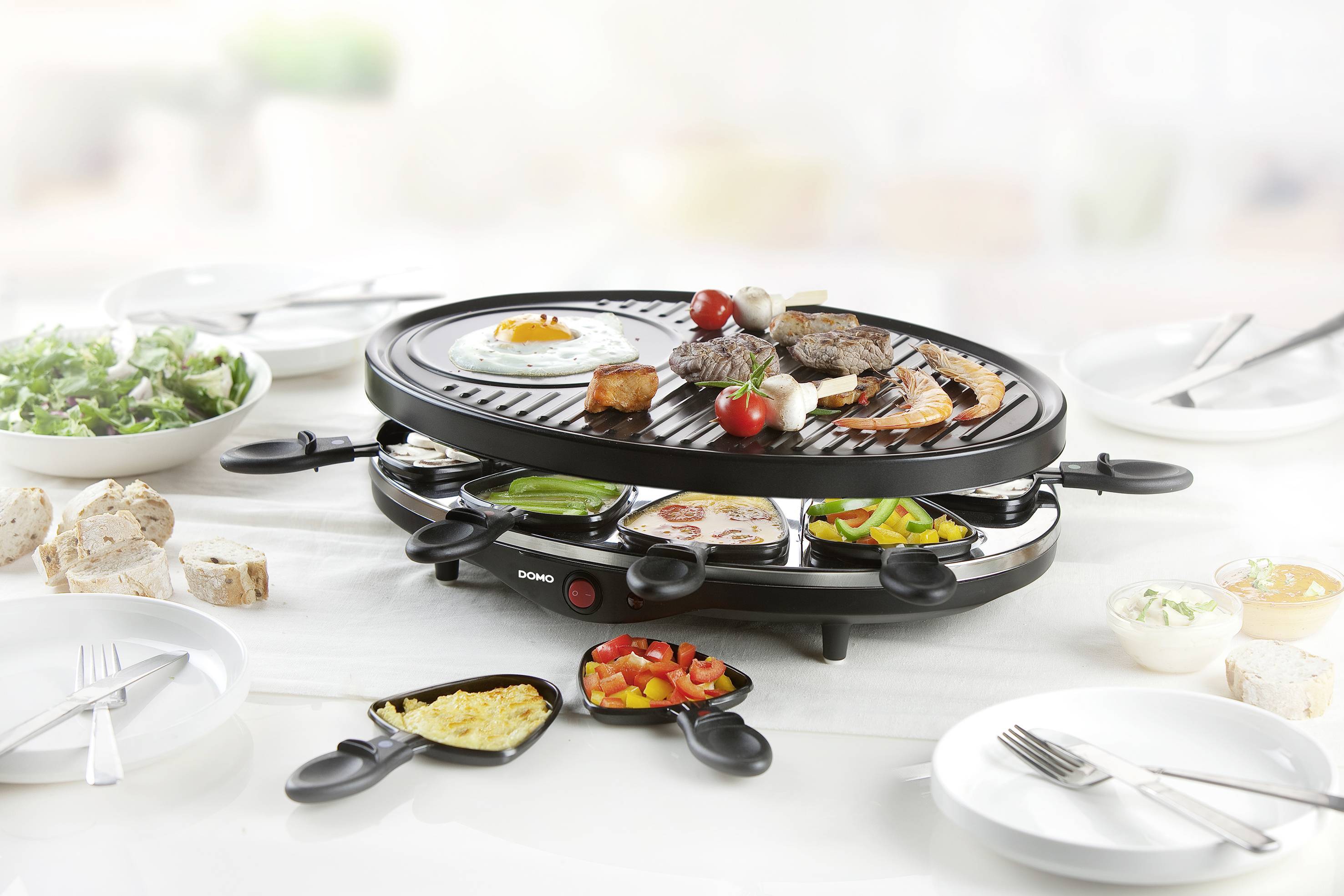 Raclette gril s rôznymi potravinami ako sú mäso, zelenina a vajce, obklopený miskami s chlebom, šalátom a omáčkou.