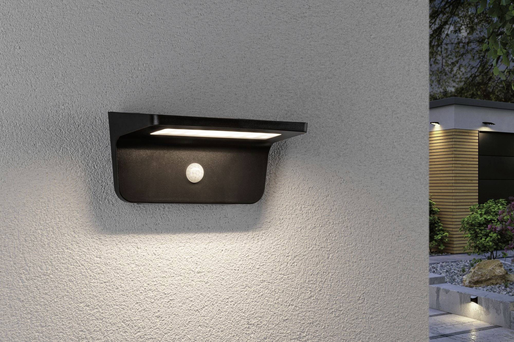 Vonkajšie svietidlo s integrovaným pohybovým senzorom osvetľuje fasádu domu, pričom záhrada v pozadí je tiež osvetlená.