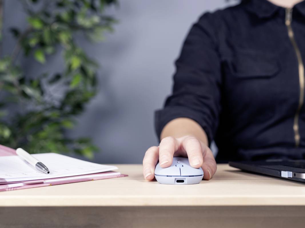 Osoba sedí pri stole a drží počítačovú myš. Vedľa nej ležia notes s perom a laptop.