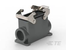 Lesklá, kovová strojová súčiastka s dvoma upevňovacími ramenami. V pozadí rozmazaný nápis značky.