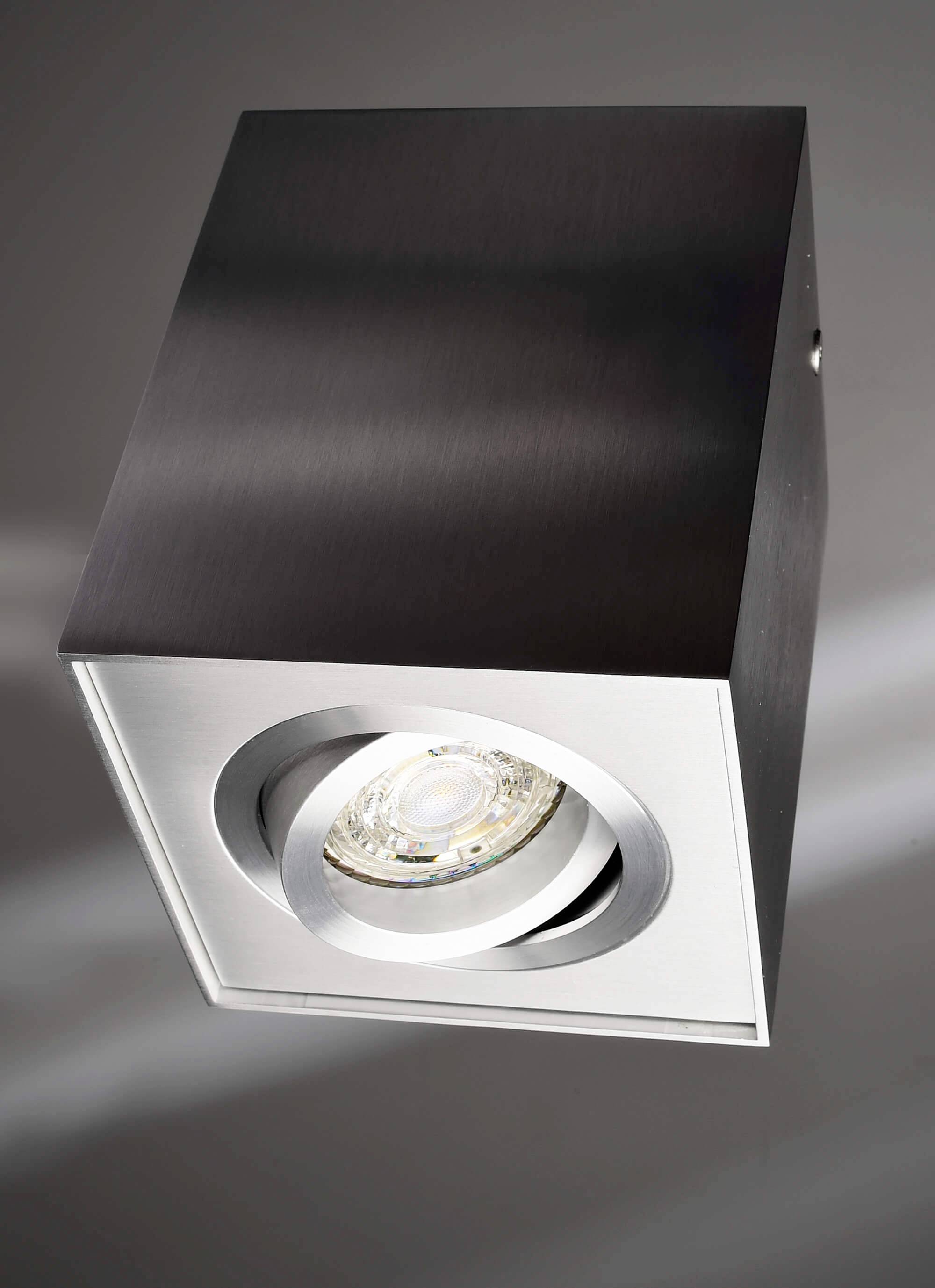 Stropný bodový svetlomet v tvare kocky s kovovým povrchom. Svietidlo smeruje svetlo smerom nadol a je navrhnuté v jednoduchom, modernom štýle.