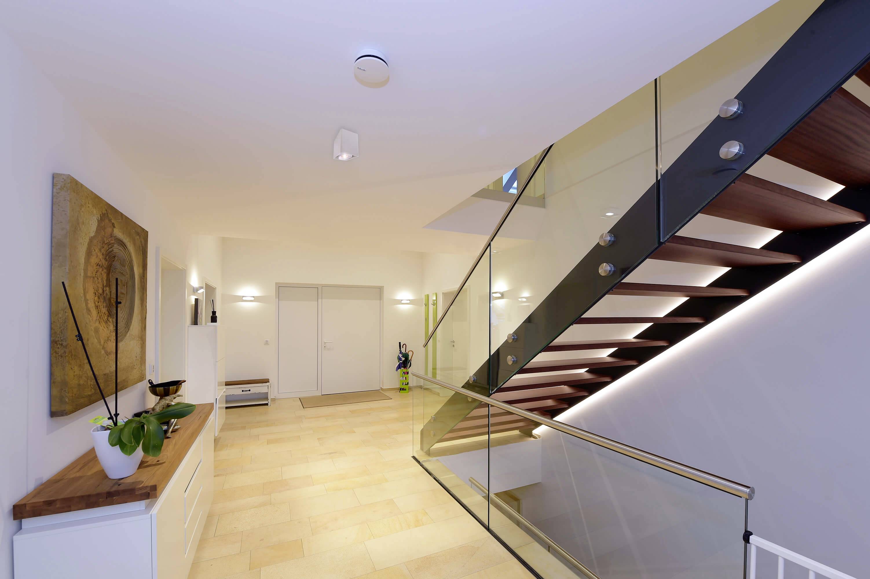 Obytný priestor s krémovou dlažbou, moderným schodiskom z tmavého dreva a skleneným zábradlím. Na stene visí abstraktné umelecké dielo.