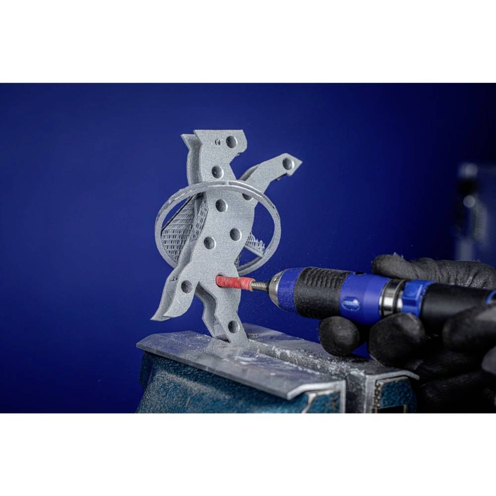 'Niekto drží červeno-čierno-šedý nástroj a opracúva strieborný kovový diel na zveráku pred modrým pozadím.'