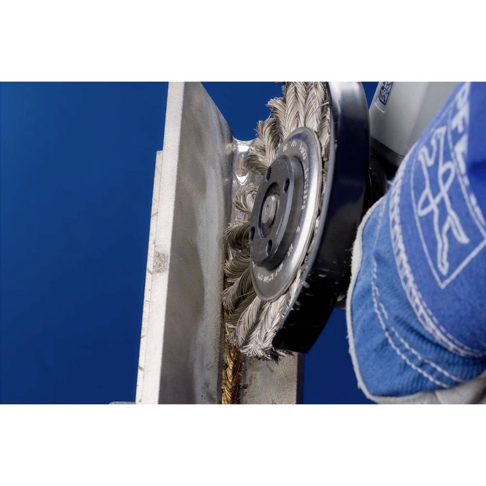 Drôtená kefa čistí hranu kovovej dosky. Je vidieť modré pozadie. Pracovník má oblečený modrý ochranný odev.