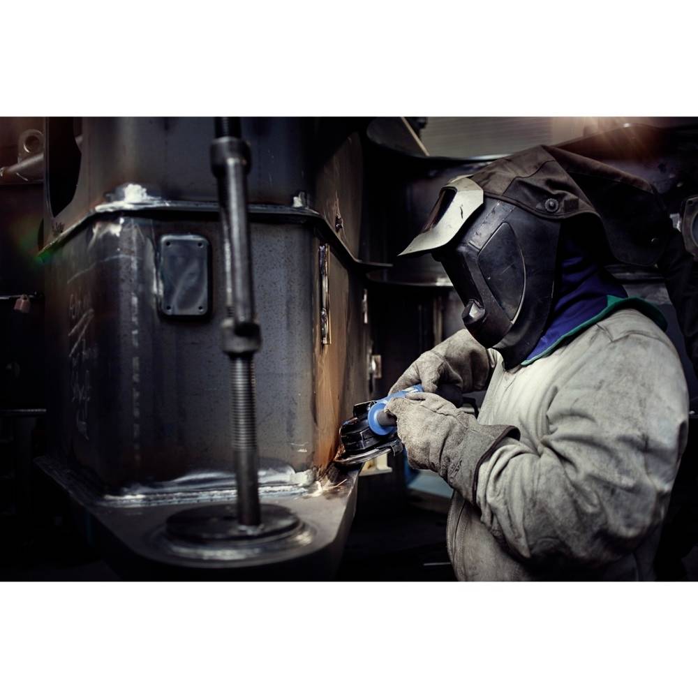 Zvárač v ochrannom odeve sa sústredeným pohľadom venuje kovovému dielu. Okolité detaily sú nejasné, zváracie zariadenie je viditeľné.