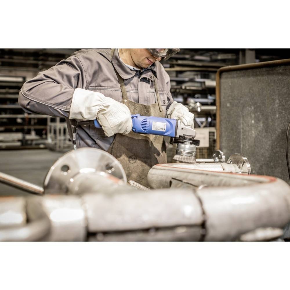 Robotník v ochrannom odeve brúsi kovové potrubie modrým uhlovou brúskou v dielni obklopenej potrubím.