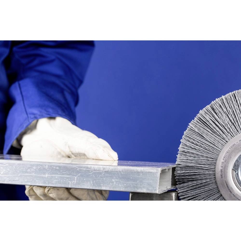 Osoba v modrom pracovnom oblečení drží kovový kus vedľa elektrickej brúsnej kotúča pripraveného na brúsenie.