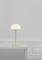 Minimalistická stolná lampa so zlatým stojanom a bielym tienidlom stojí na jednoduchom bielom stole pred sivou stenou.