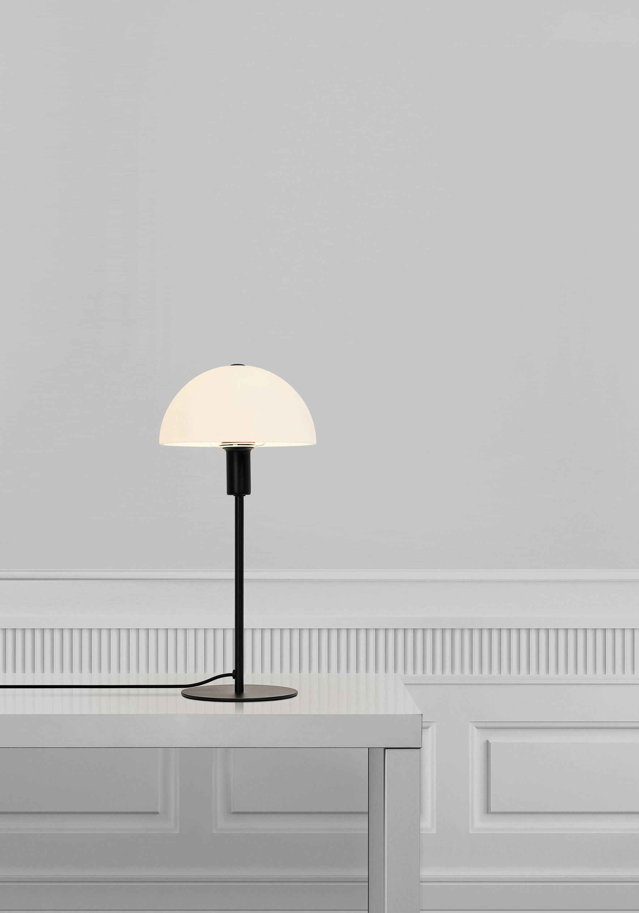 Minimalistická stolná lampa s okrúhlym bielym tienidlom stojí na bielom stole pred svetlosivou, jednoduchou stenou.
