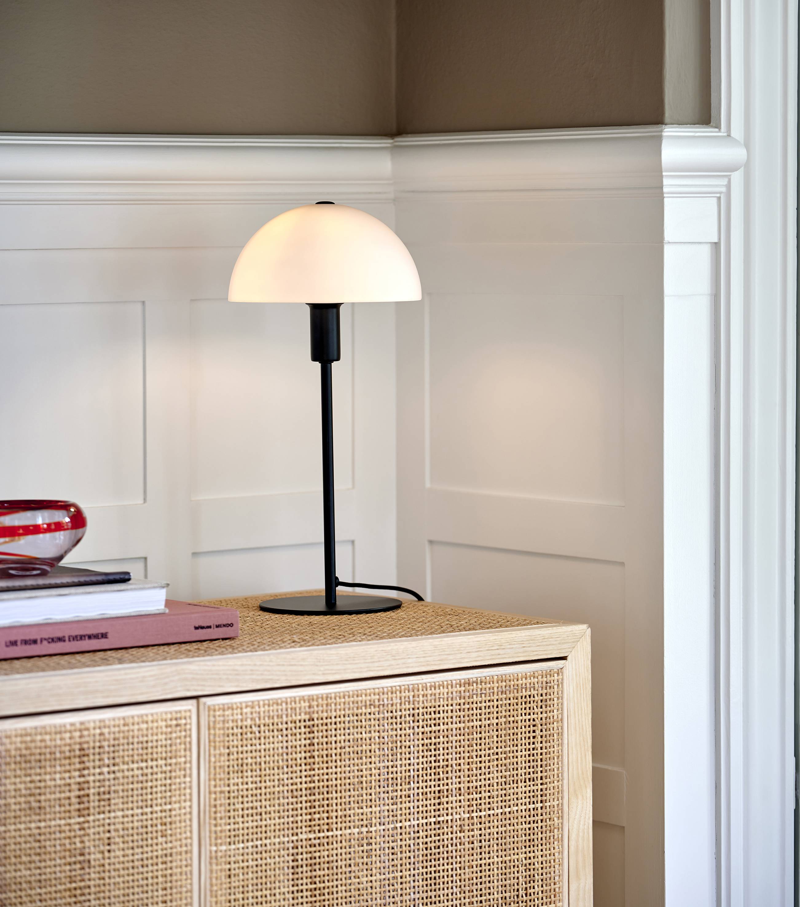 Stolová lampa s polkruhovým tienidlom stojí na komode. Vedľa nej ležia knihy a červená sklenená dekorácia.