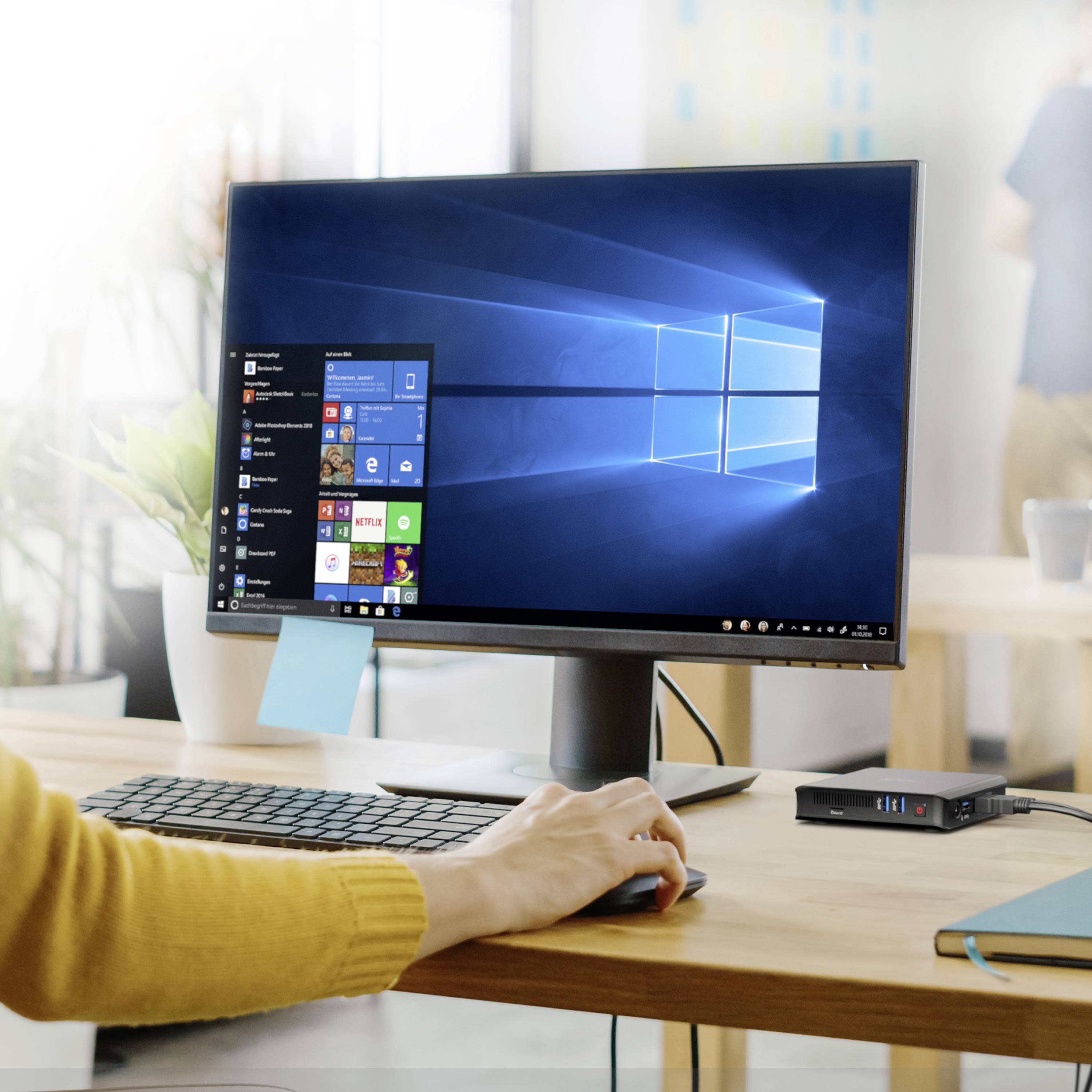 Počítačový monitor zobrazuje pracovnú plochu systému Windows 10. Na stole sa nachádza klávesnica a myš. V pozadí je nejasne vidieť osobu.