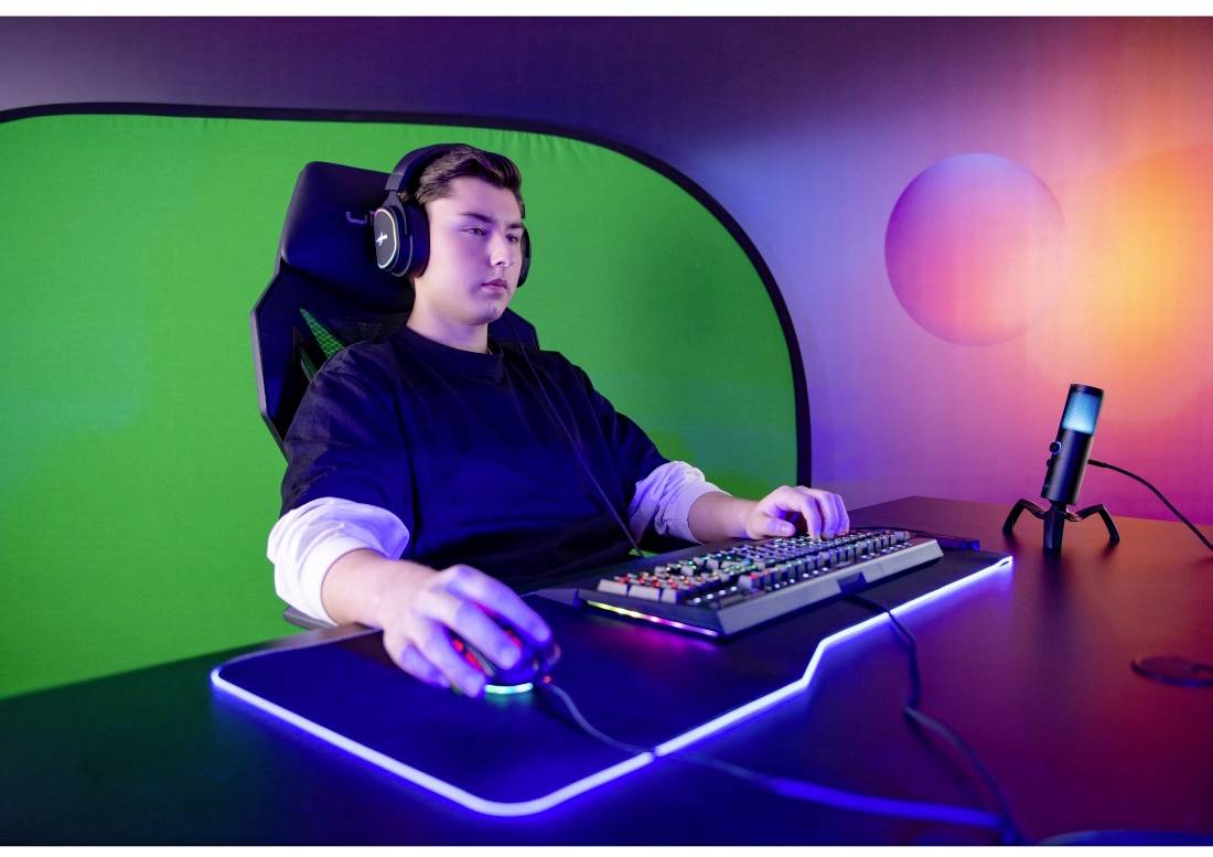 Mladý človek so slúchadlami sedí pri stole s klávesnicou a myšou, pred zeleným pozadím, osvetlený farebnými svetlami.