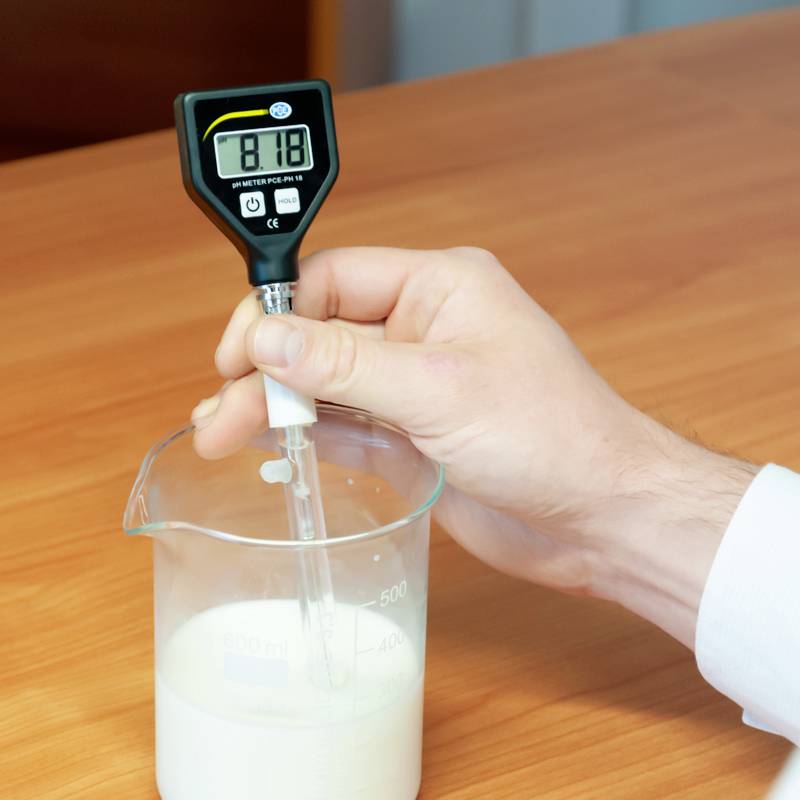 Jedna ruka drží pH meter, ktorý je ponorený do kvapaliny v odmernom valci. Na displeji je zobrazená hodnota 'pH 6,60'.