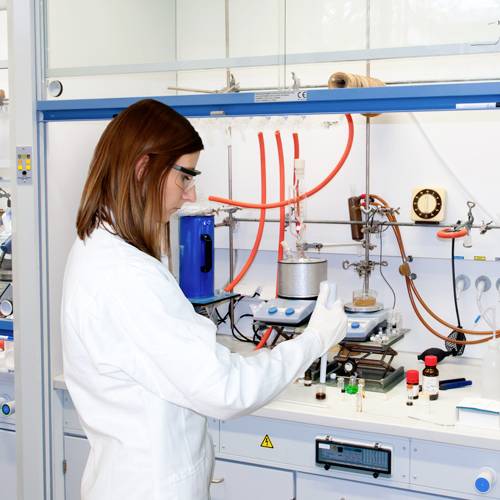 Vedkyňa pracuje v laboratóriu. Má na sebe biely laboratórny plášť a vykonáva experiment s chemickými látkami.