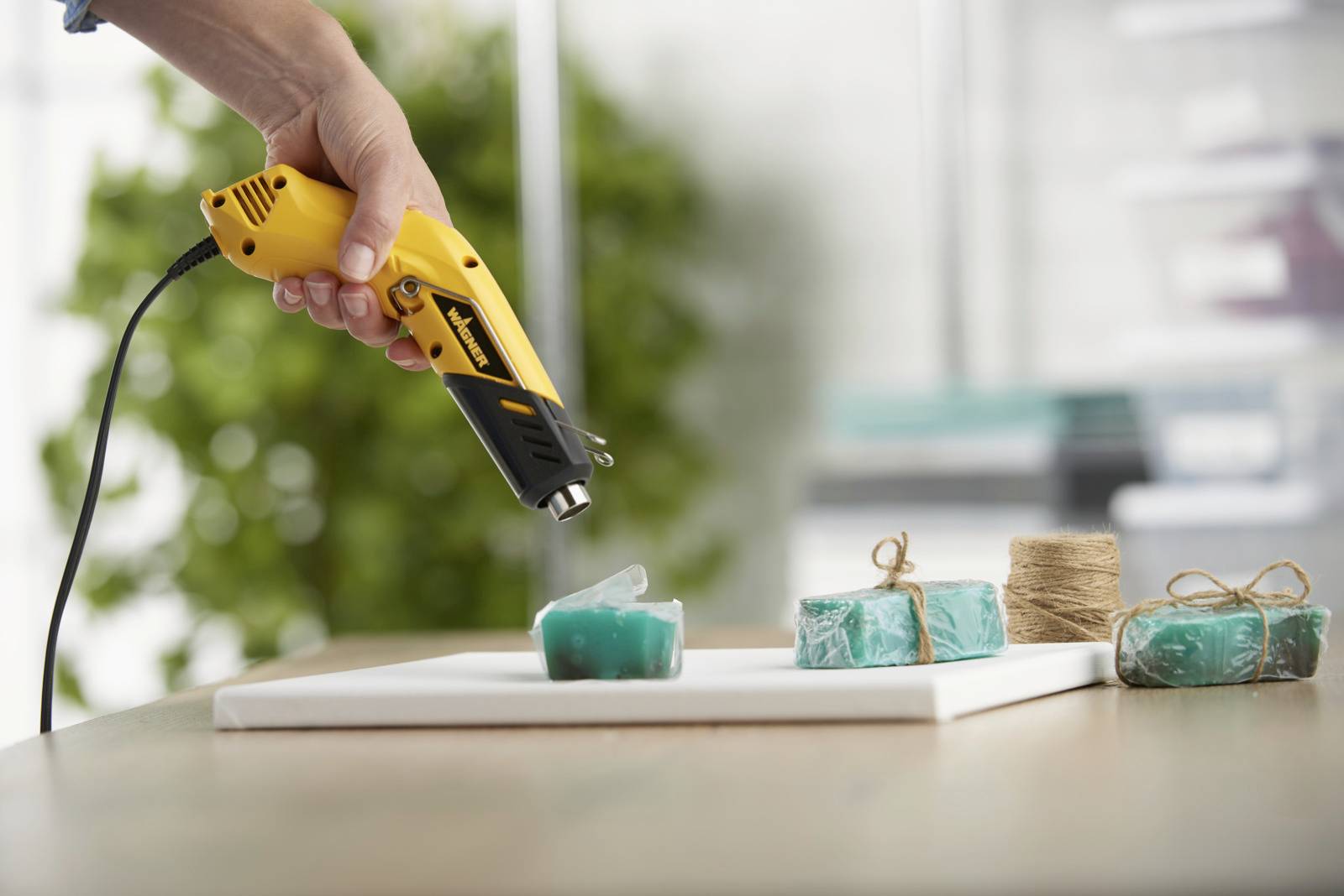 Jedna ruka drží teplovzdušnú pištoľ nad niekoľkými zelenými kúskami mydla, ktoré ležia na stole. Vedľa leží bavlnená šnúra.