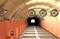 Cestný tunel s núdzovým telefónom osvetleným na ľavej stene a ventilátormi na strope. Svetlo na konci tunela.