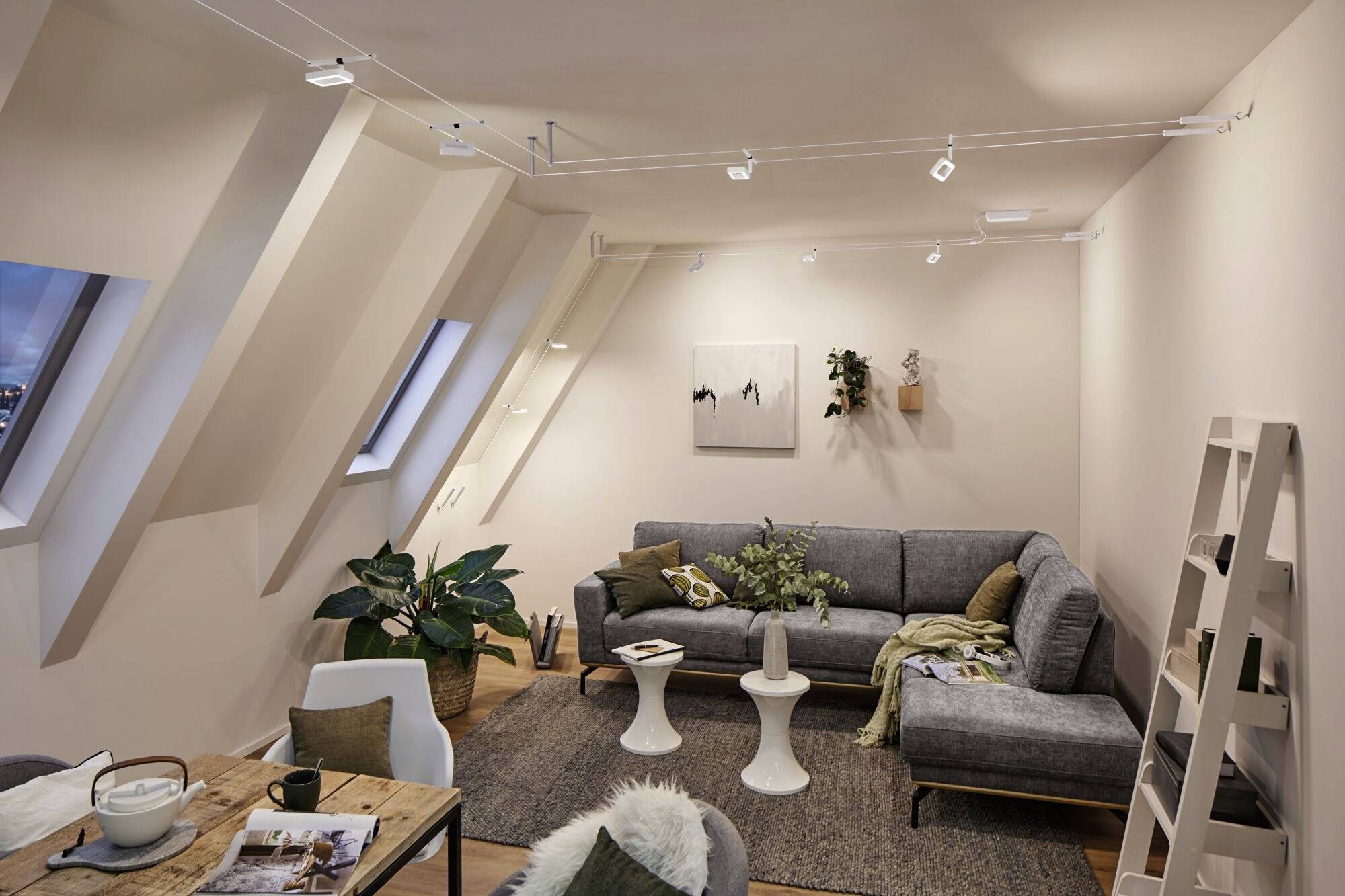 Útulná obývačka v podkrovnom byte so sivou rohovou sedačkou, konferenčným stolíkom, rastlinami a umeleckými dielami na stene. Teplé svetlo.