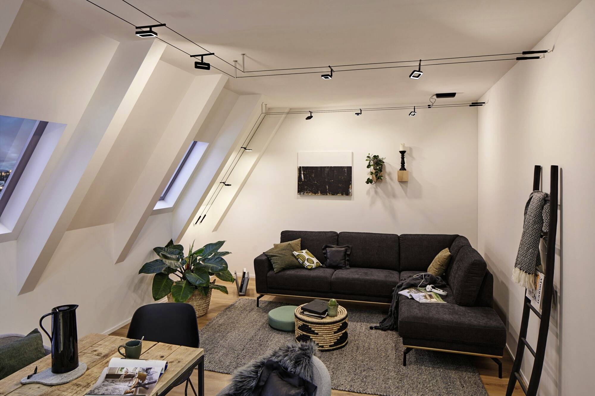 Útulná obývačka v podkroví so šikmými stenami, sivou sedačkou, dreveným stolom a rastlinami. Moderné osvetlenie na strope.