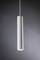 Štíhla, valcovitá závesná lampa z kovu, ktorá visí v modernom, neutrálnom priestore. Svieti smerom nadol.