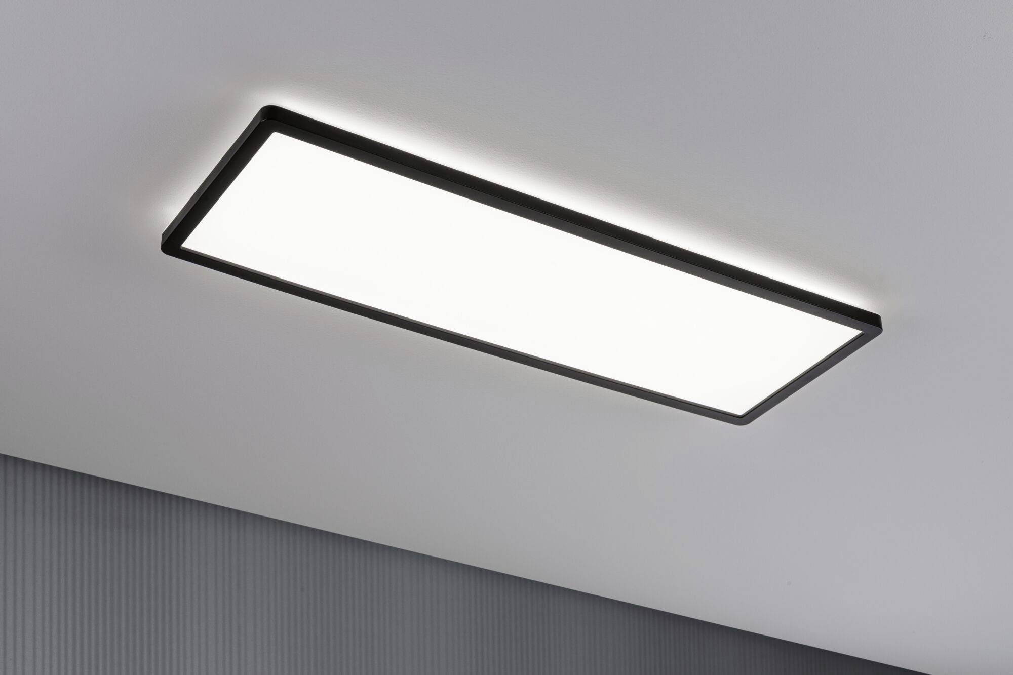 Obdĺžniková stropná lampa s čiernym rámom na bielom strope. Vyžaruje jasné, rovnomerné svetlo.