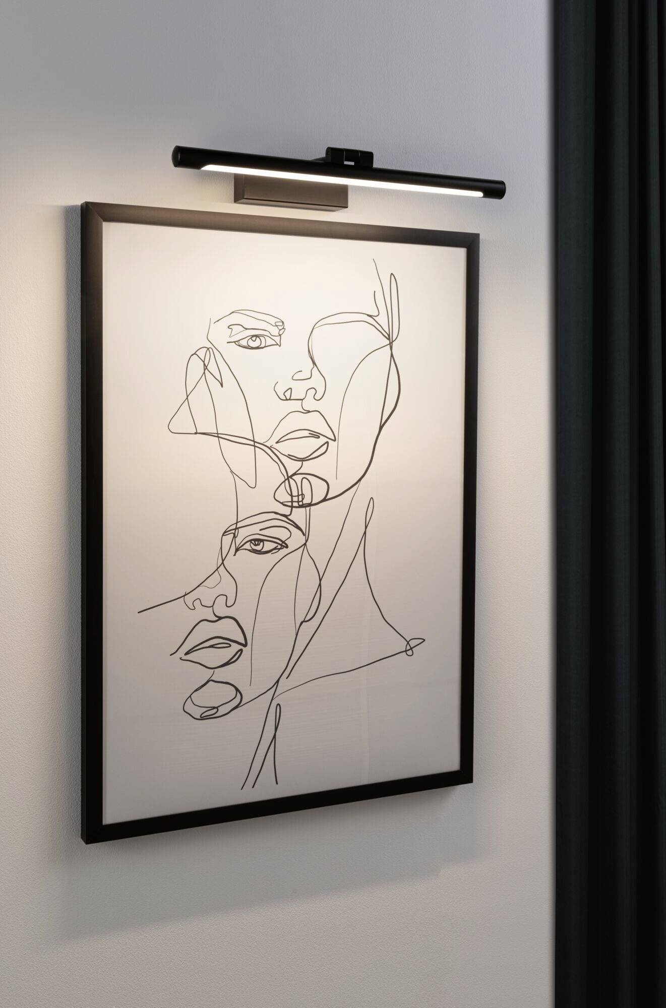 Na stene visí obraz, ktorý zobrazuje minimalistický portrét s viacerými tvárami nakreslenými jedinou neprerušenou líniou.