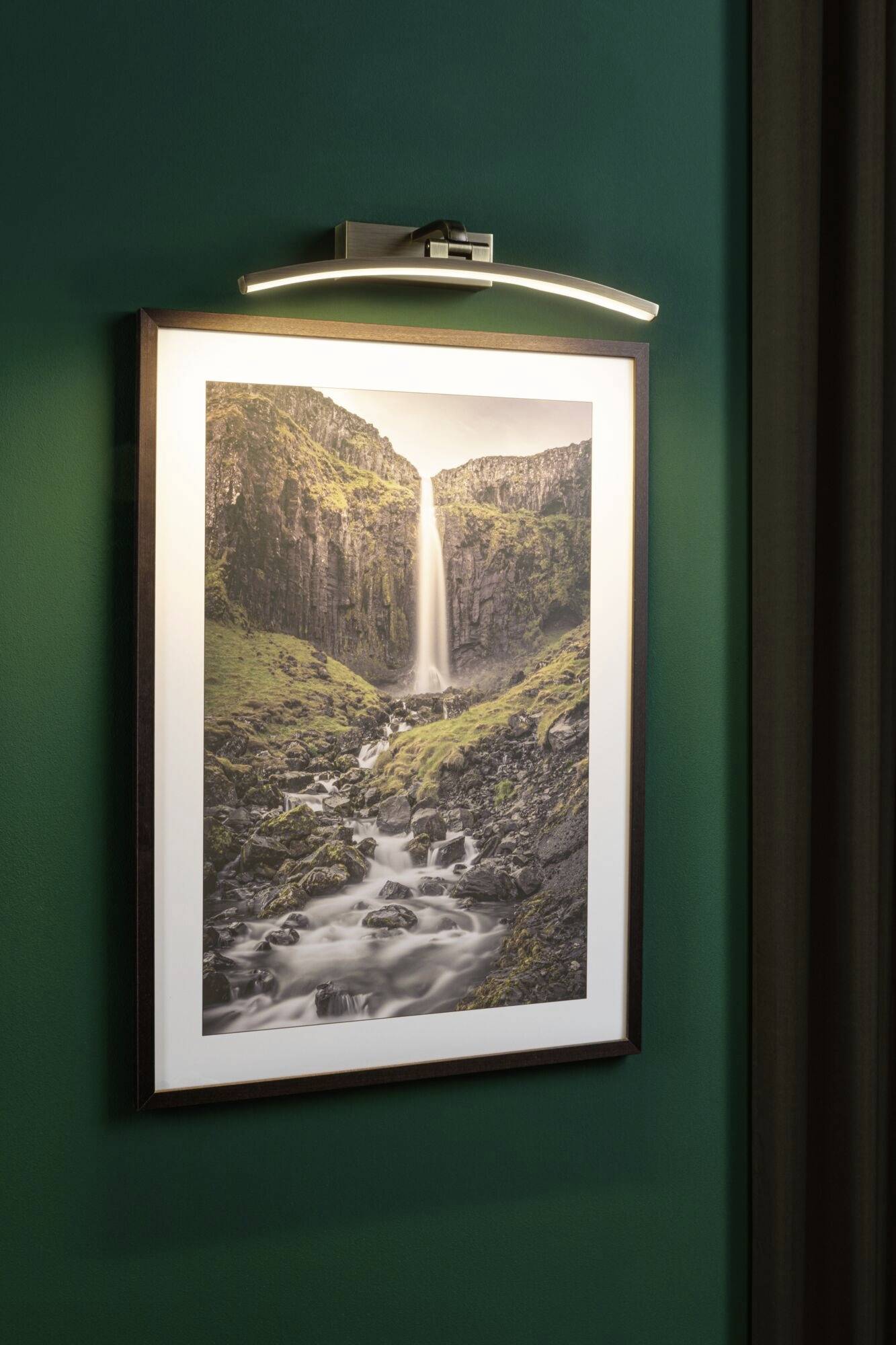 Na tmavozelenej stene visí osvetená fotografia vodopádu v hornatej krajine.