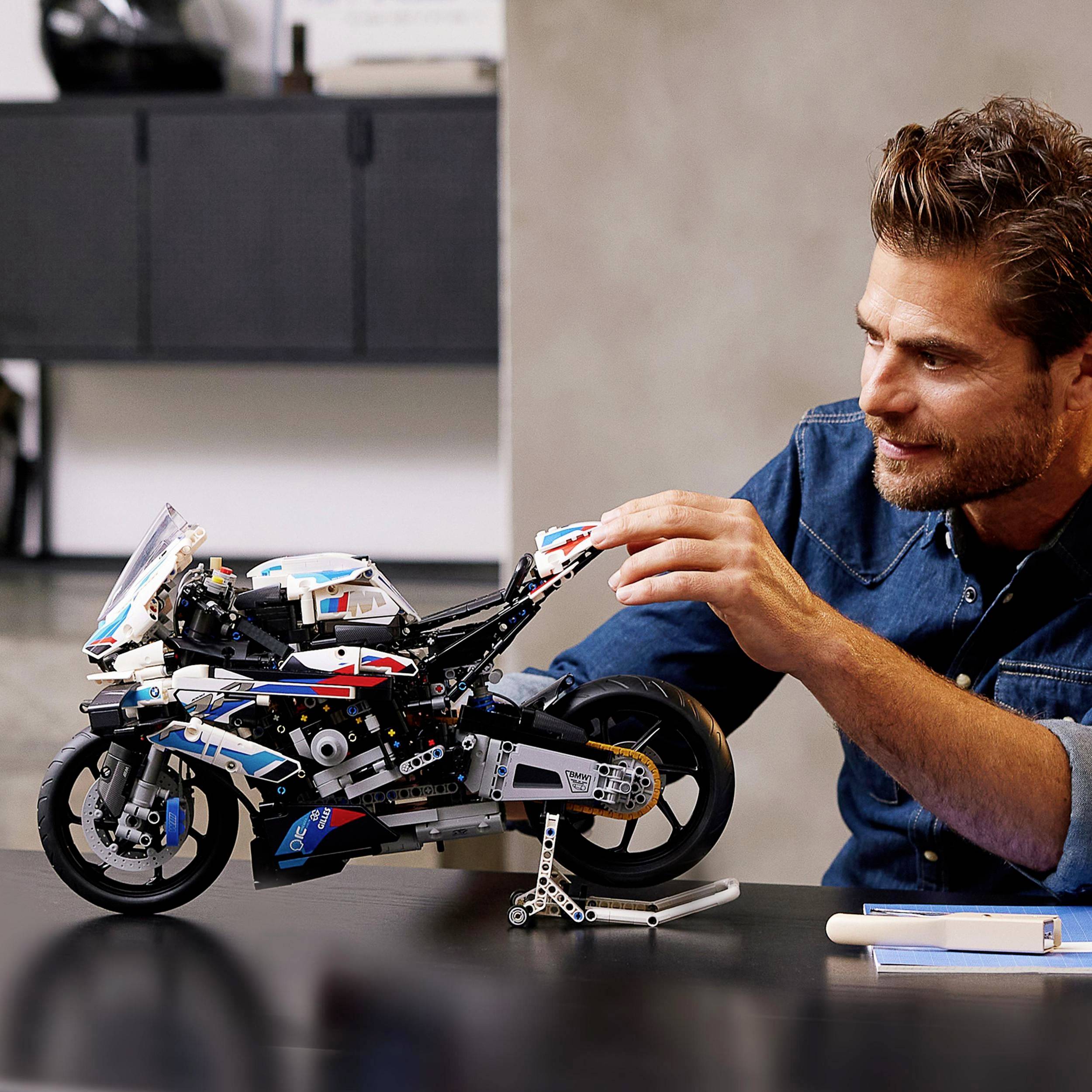 Usmiaty muž skladá model motorky z kociek na stole. V pozadí sú rozmazané kusy nábytku.