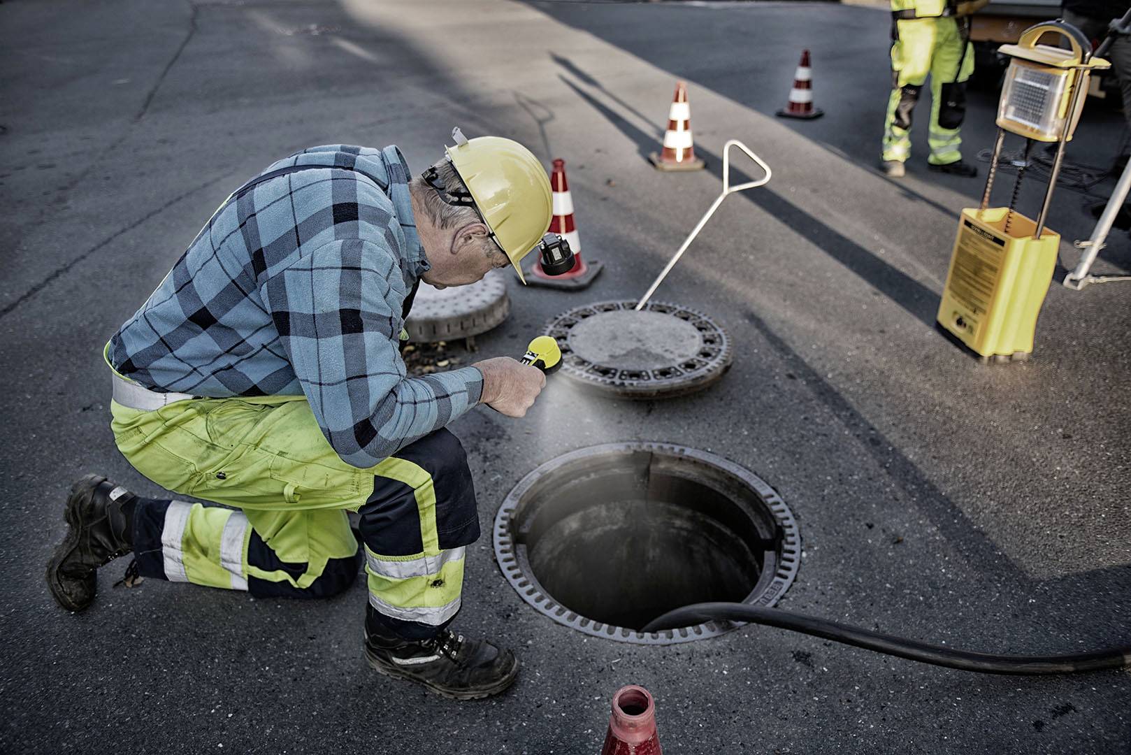 Robotník v ochrannom oblečení kontroluje svetlom baterky otvor kanálového poklopu na ceste, obklopený výstražnými kužeľmi.
